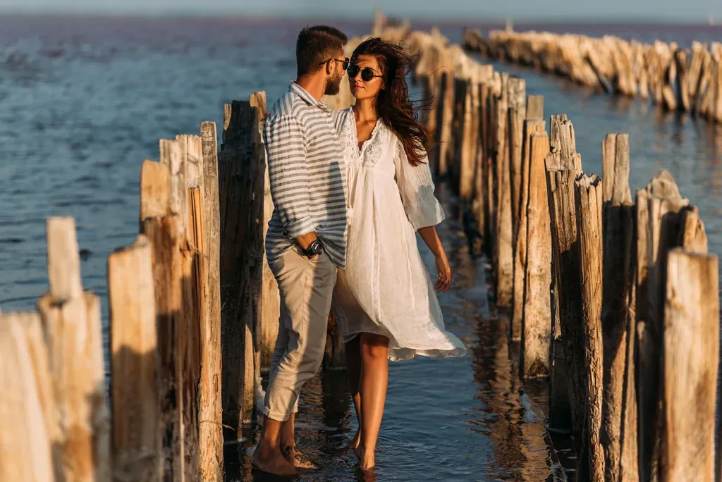 Couple enlacé marchant pieds nus le long d'une jetée en bois au bord de la mer au coucher du soleil