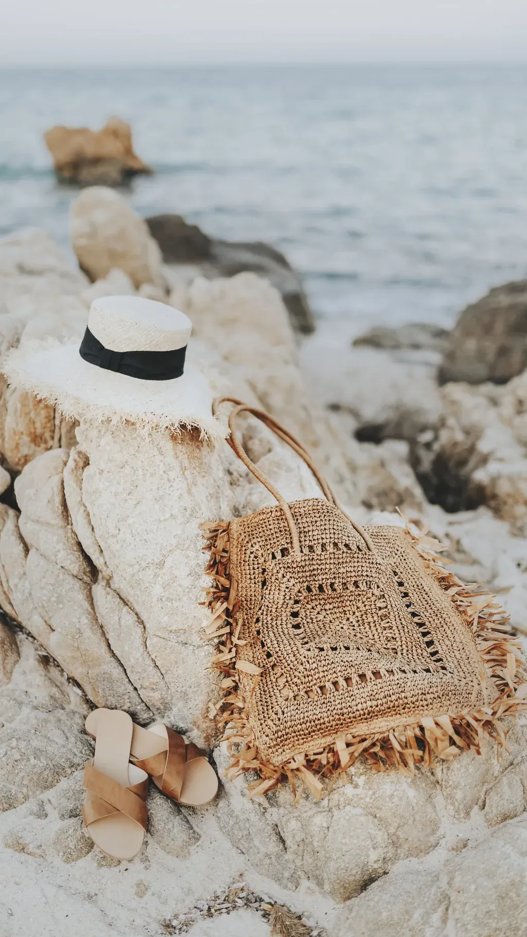 Sac en crochet couleur naturelle et chapeau de paille posés sur des rochers au bord de la mer
